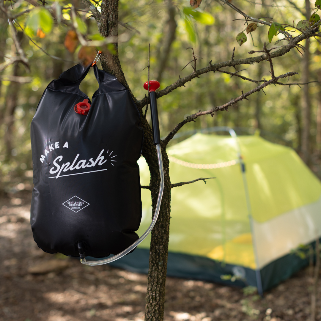 A Portable Solar Shower hangs from a tree branch, ready for use on your camping trip, with a yellow tent in the wooded background.
