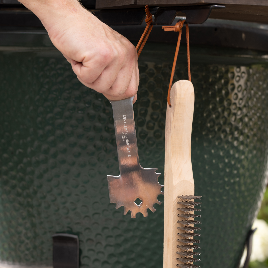 A hand holds the BBQ Care Kit Grill Scraper, hanging next to its wooden wire brush beside a green outdoor grill.