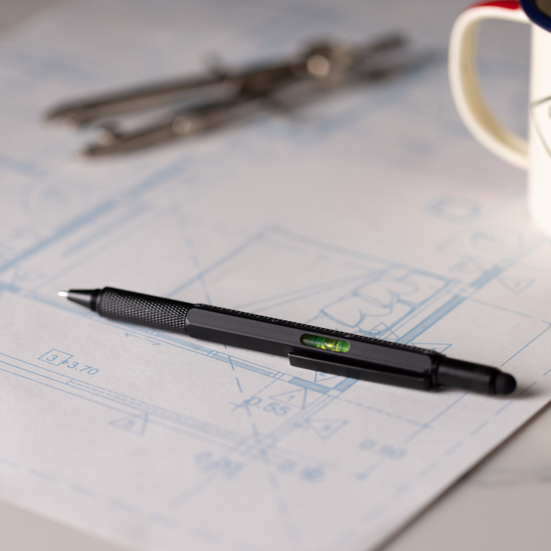 A Tooling Pen sits atop an architectural blueprint, with a drafting compass and mug nearby.