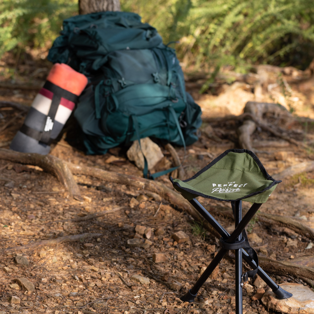The Tripod Camp Stool, a small green compact seat with a folding tripod base, sits on dirt near a large green backpack and a rolled sleeping pad in the woods.