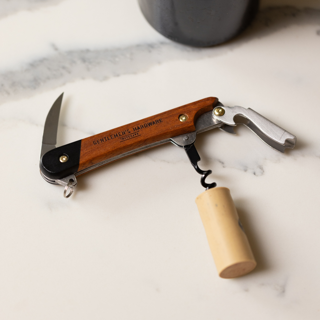 The Waiters Friend is a wooden-handled corkscrew and bottle opener with GENTLEMENS HARDWARE engraved, shown holding a cork on a light marble surface.