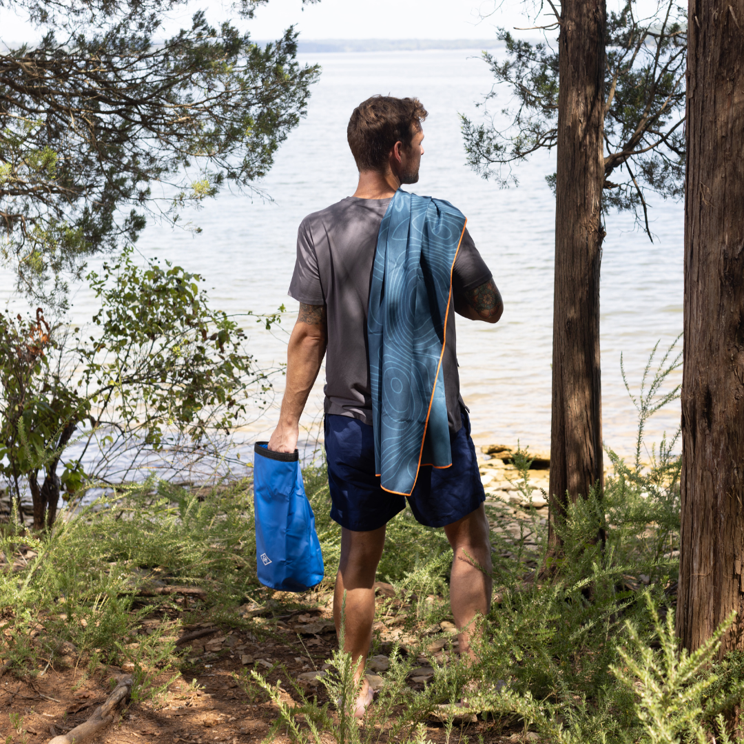 A man stands on a wooded lakeshore, looking toward the water with a blue bag in one hand and the Travel Towel draped over his shoulder.