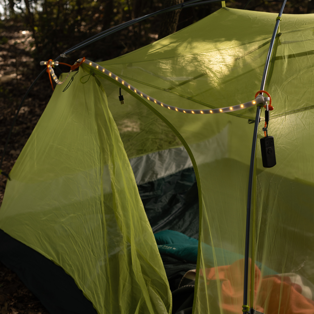 A green camping tent with LED Rope Lights attached to the frame, set up outdoors in a wooded area for convenient campsite lighting.
