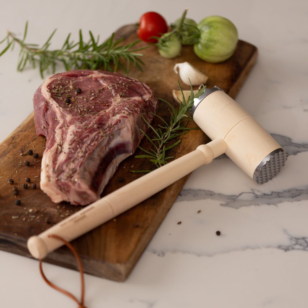 A raw steak on a wooden board with the BBQ Lovers Kit Meat Tenderizer & Thermometer, rosemary, black pepper, garlic, a tomato, and a green tomato sits on a marble countertop.