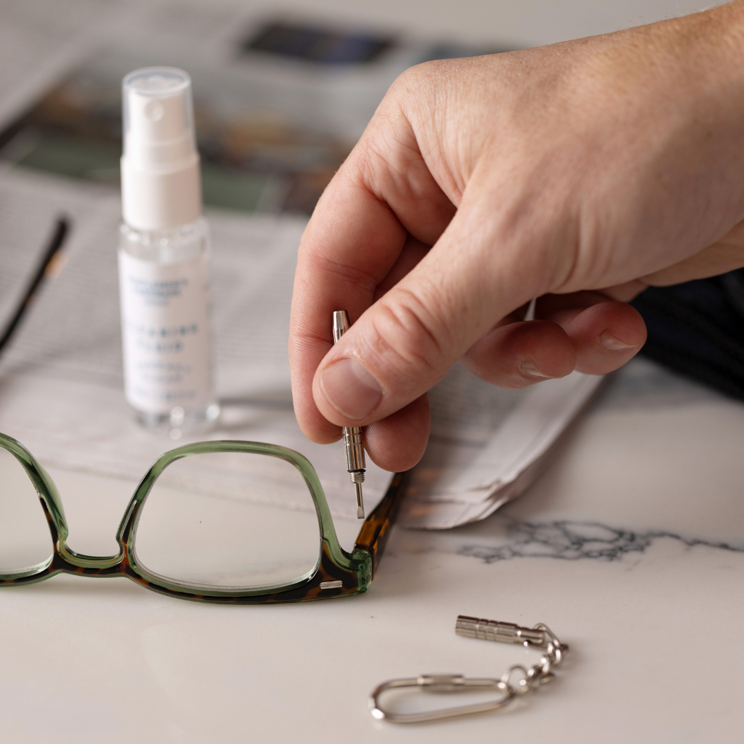 A hand tightens the hinge of green eyeglasses with a mini screwdriver from the “Squeaky Clean” Clean & Repair Kit, on a table with cleaning fluid, a keychain tool, and a newspaper.