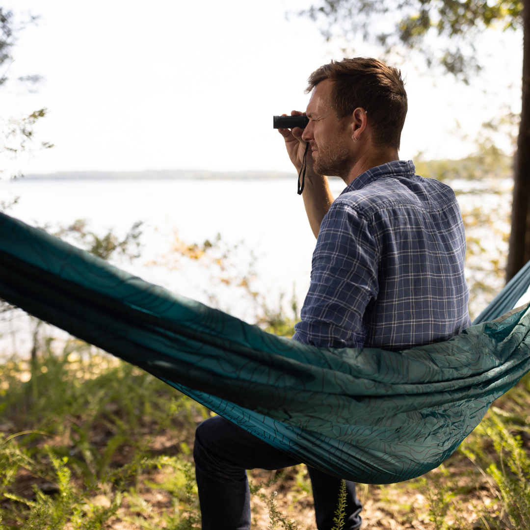 A man relaxes in a hammock by a lakeside, surrounded by trees, using his Explorer Kit to scan the water—embracing outdoor adventure in uncharted terrain.