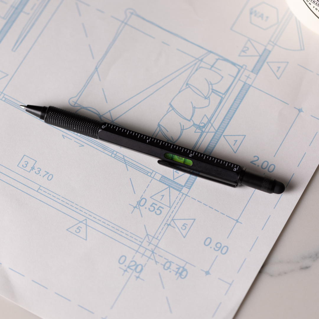 A black Tooling Pen with 6-in-1 functions and a built-in level lies on technical blueprints displaying architectural drawings and measurements.