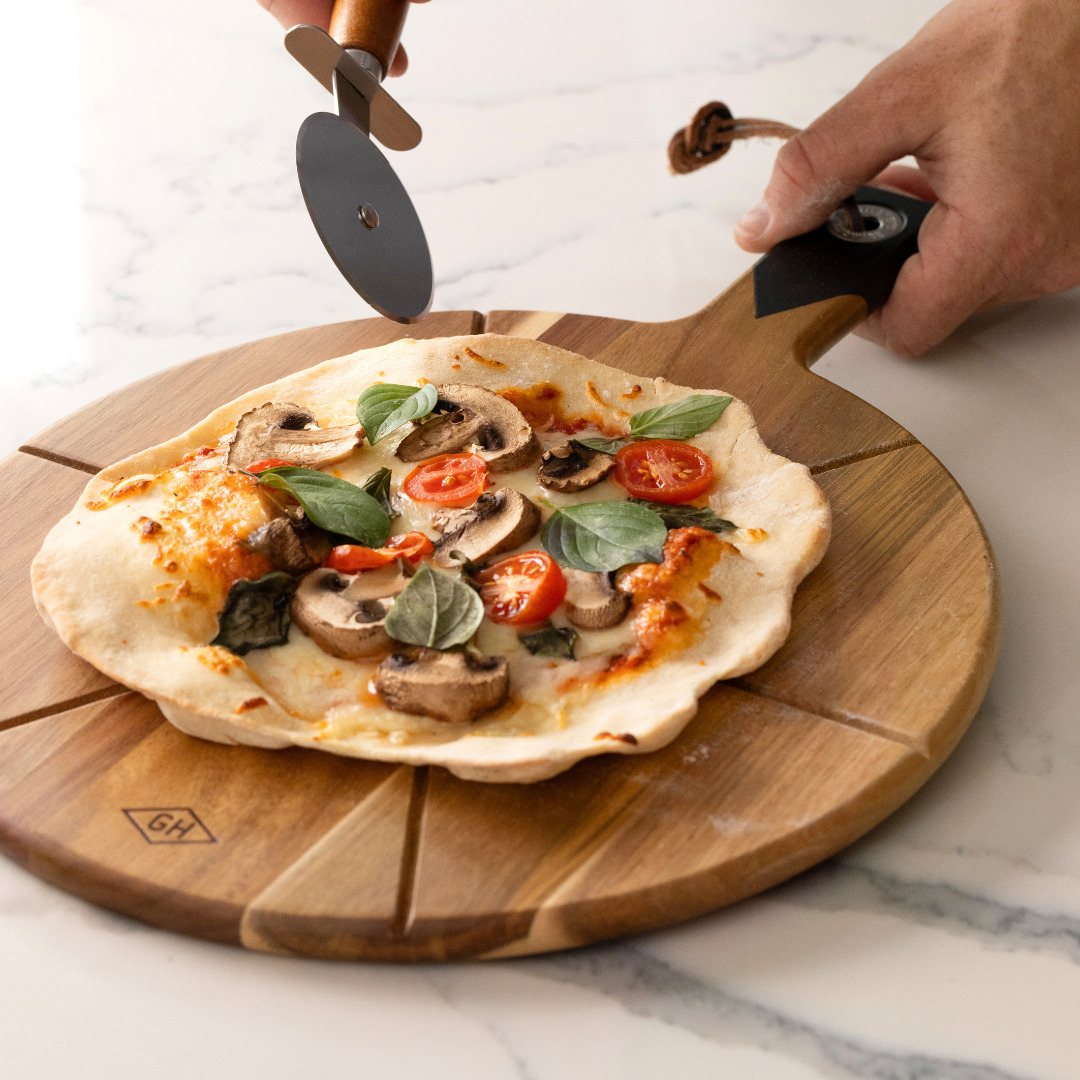 A hand slices a homemade pizza topped with mushrooms, cherry tomatoes, and basil using the Pizza Board with pizza cutter.