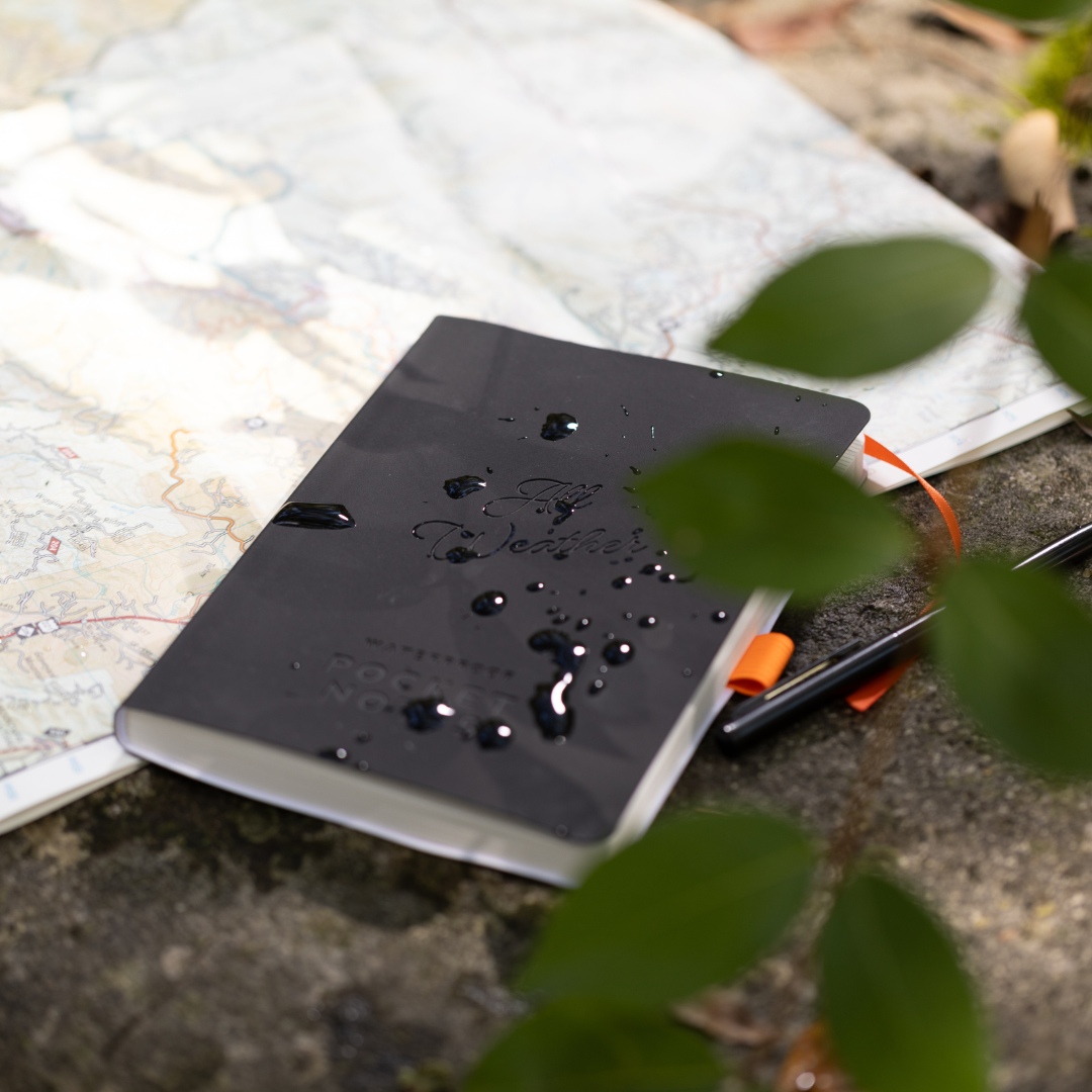 A black Waterproof Notebook rests on an outdoor map, its stonepaper cover beaded with water droplets and partly hidden beneath lush green leaves.