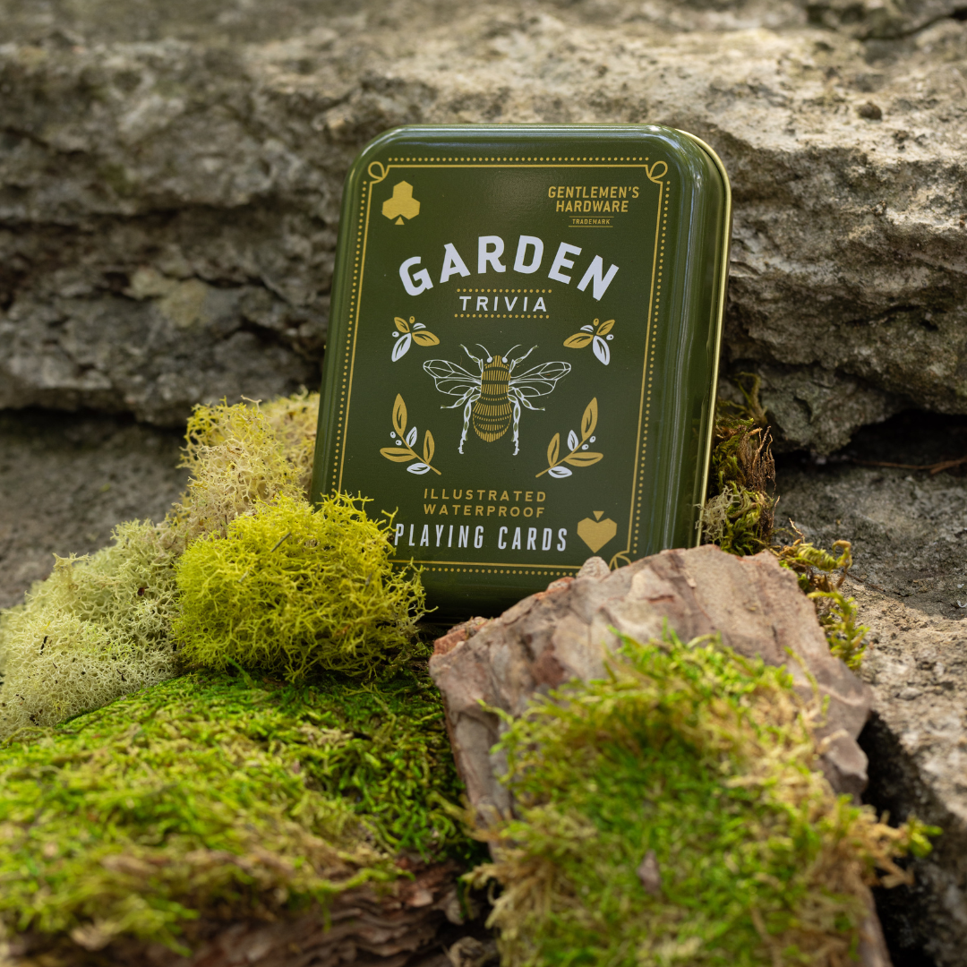 A tin of Gardeners Tips Waterproof Playing Cards, featuring garden-themed illustrations, is shown outdoors on mossy rocks and bark.