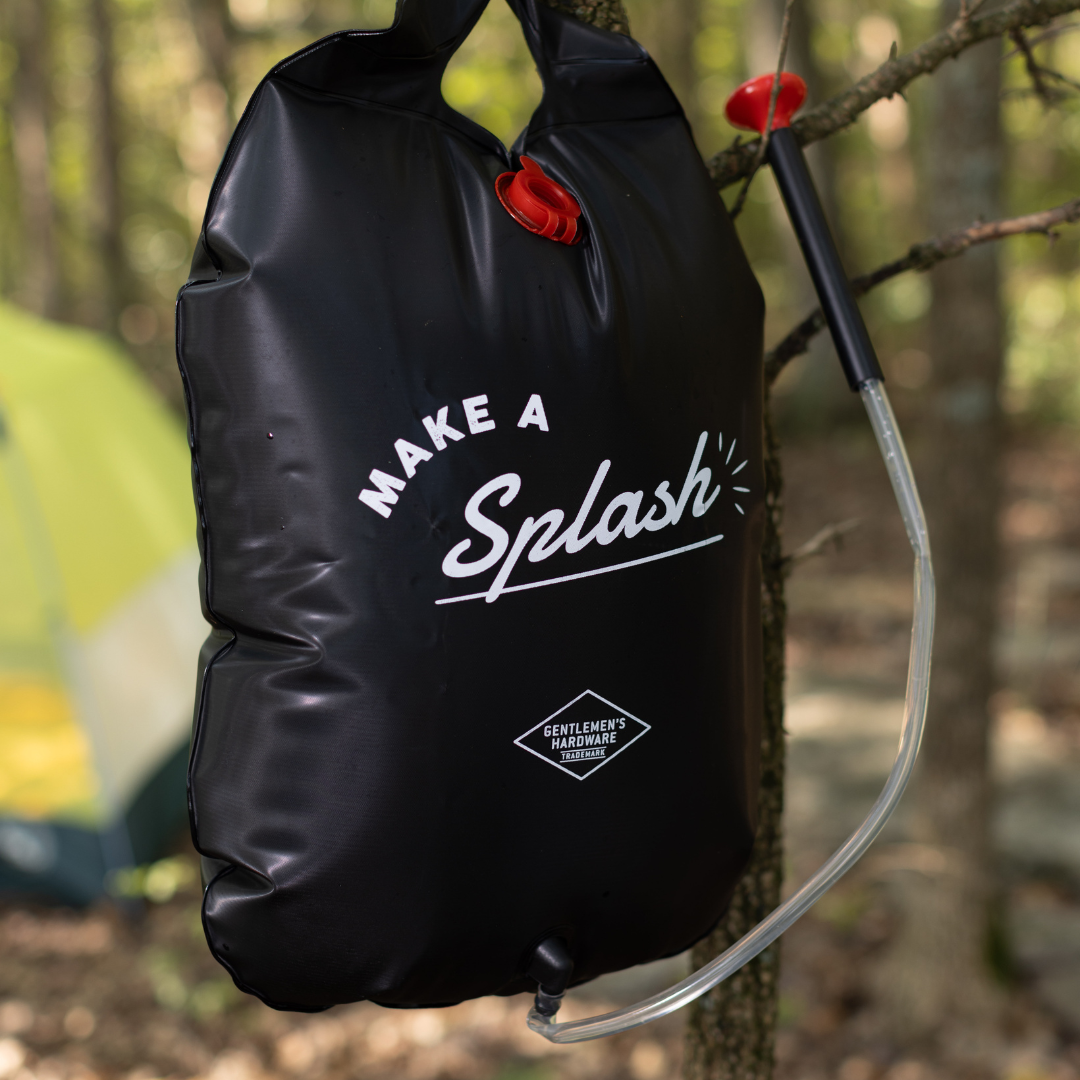 A Portable Solar Shower hangs from a tree branch with a hose attached, ready for your camping trip; a tent is visible in the background.