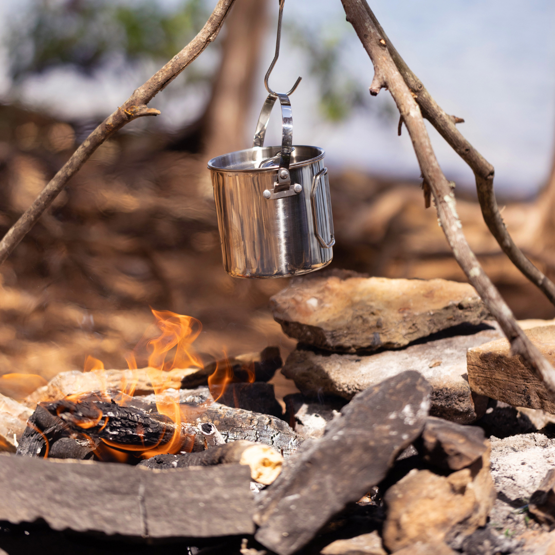 The Campfire Cooking Pot hangs from a stick over a stone-surrounded campfire with burning wood, making it ideal for outdoor wilderness cooking.