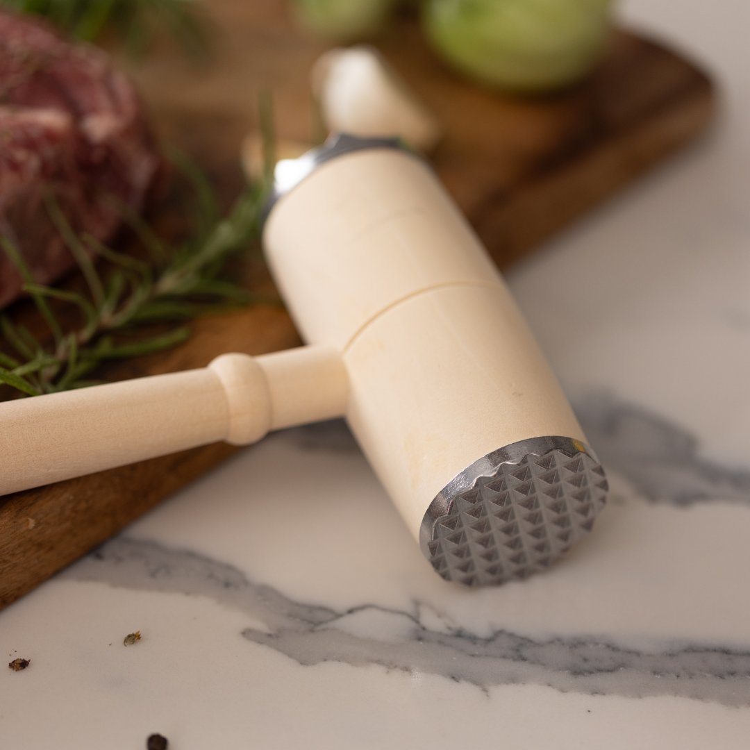 The BBQ Lovers Kit Meat Tenderizer & Thermometer, featuring a dual metal head, rests on a marble countertop beside fresh rosemary, raw meat, and green veggies—an essential toolset for every grilling enthusiast.