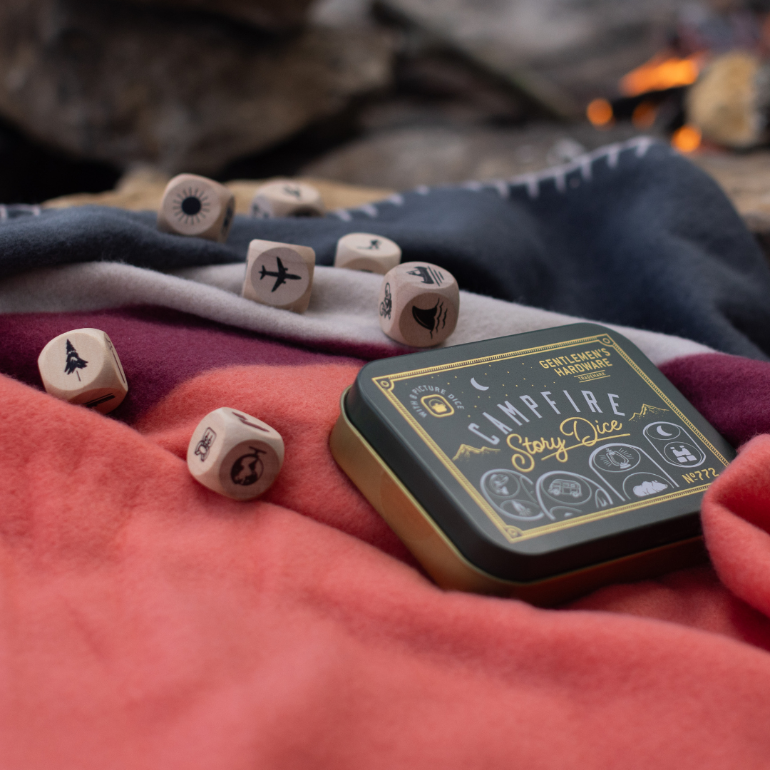 The Campfire Story Dice tin sits on a vibrant blanket outdoors, with wooden dice nearby—perfect for sparking creativity and setting the stage for an imaginative night under the stars.
