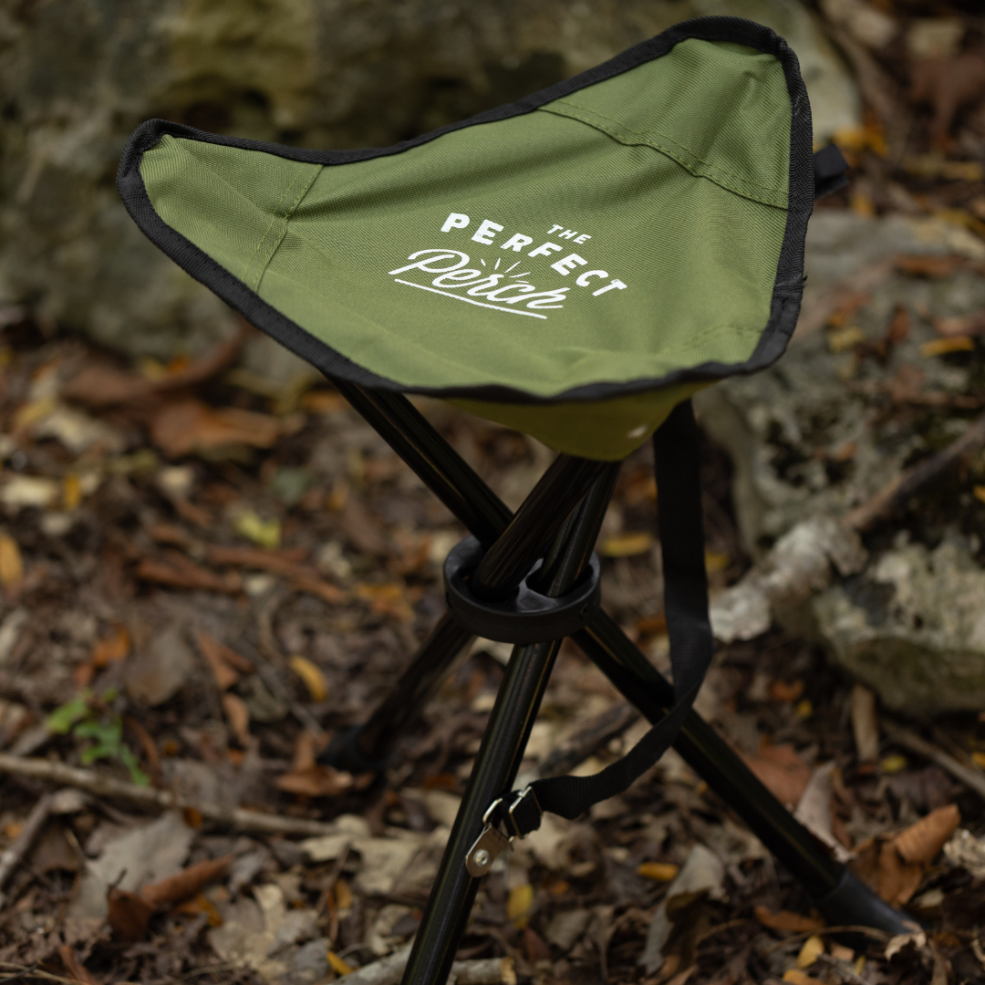 A Tripod Camp Stool with a green seat, tripod folding base, and The Perfect Rest printed on it, set outdoors on ground strewn with dry leaves and rocks.