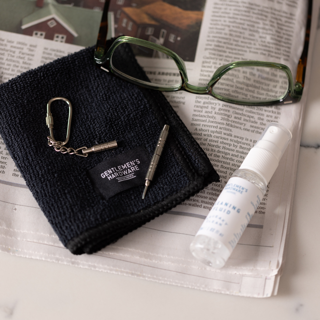 The Squeaky Clean Clean & Repair Kit, with green glasses, a black cleaning cloth, mini screwdriver keychain, and cleaning fluid spray bottle, is displayed on an open newspaper.