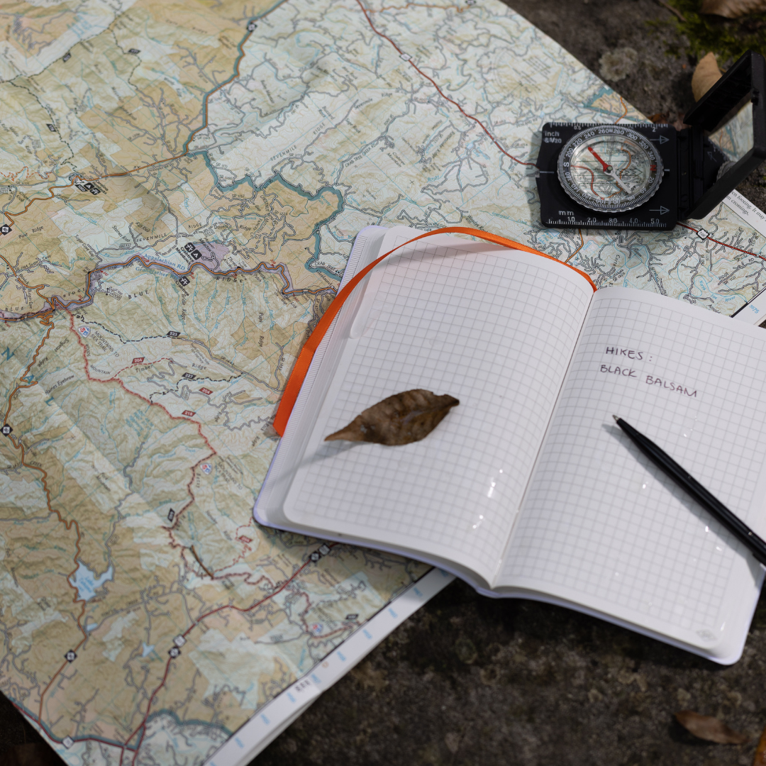 A topographic map, a compass, and an open Waterproof Notebook with a pen and a leaf are on the ground; the notebook page reads Hikes Black Balsam.