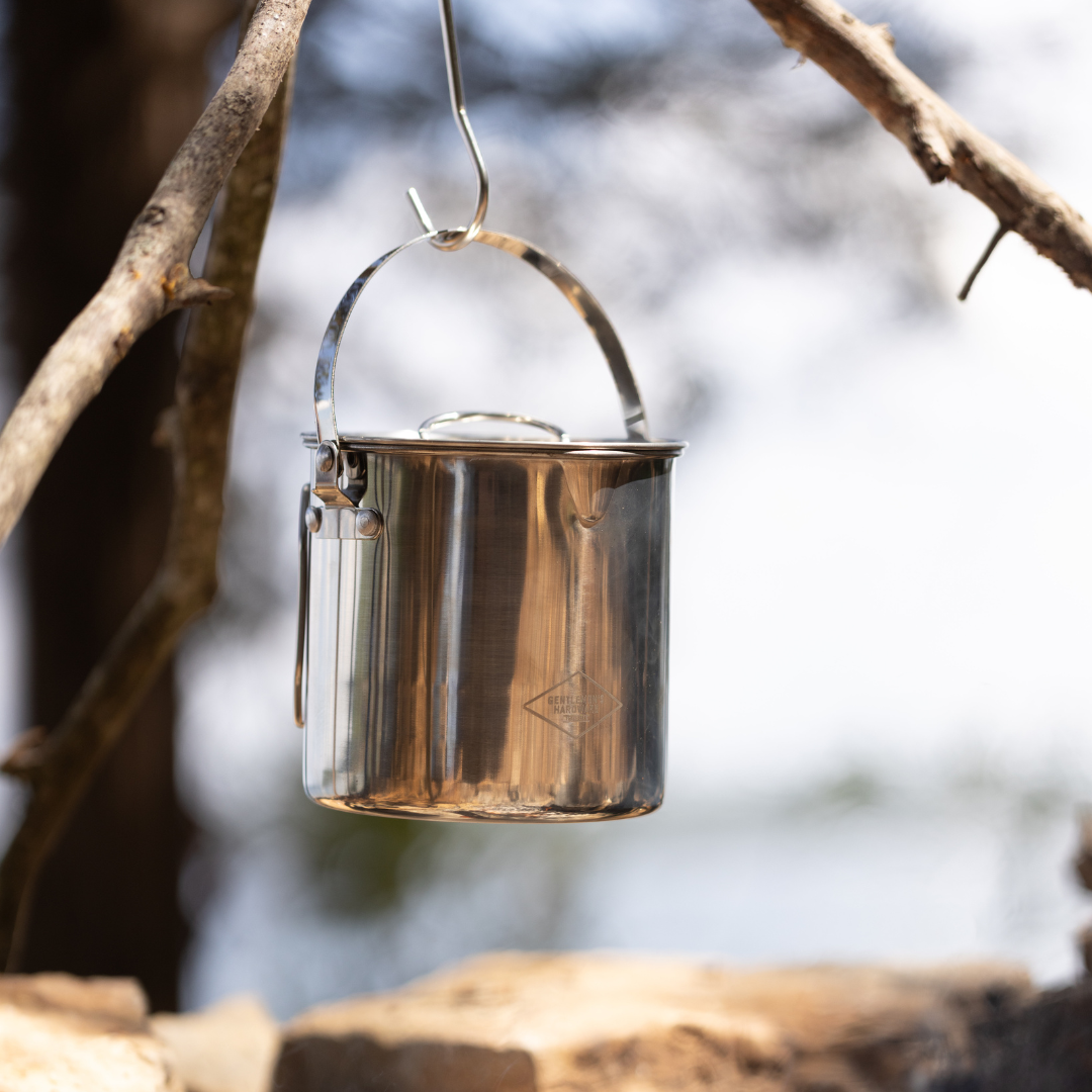 The Campfire Cooking Pot with lid hangs from a branch outdoors, ideal for wilderness cooking. Blurred trees and rocks set the scene for this essential piece of portable cookware, perfect for every adventure.