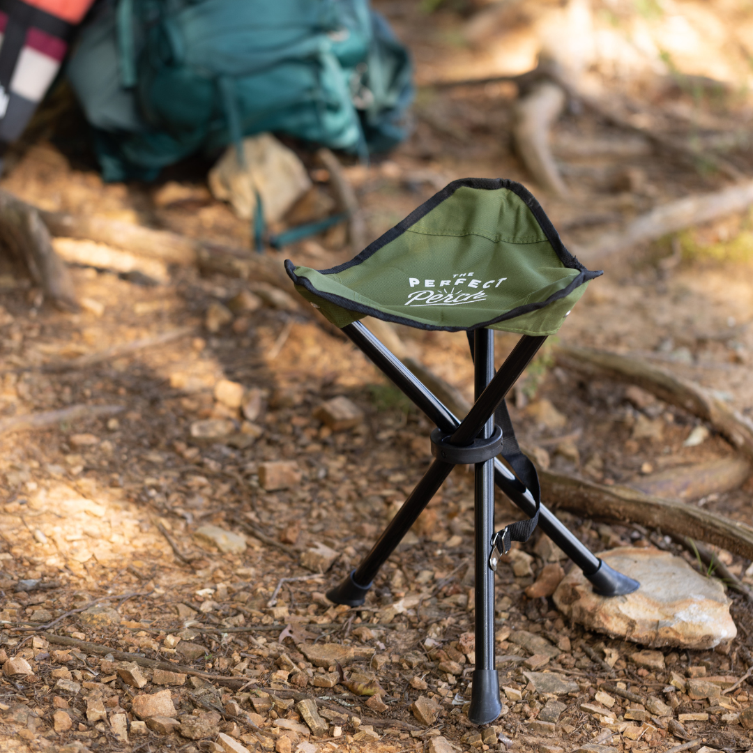 The Tripod Camp Stool is set up on rocky ground outdoors, with a green backpack and camping gear visible in the background.