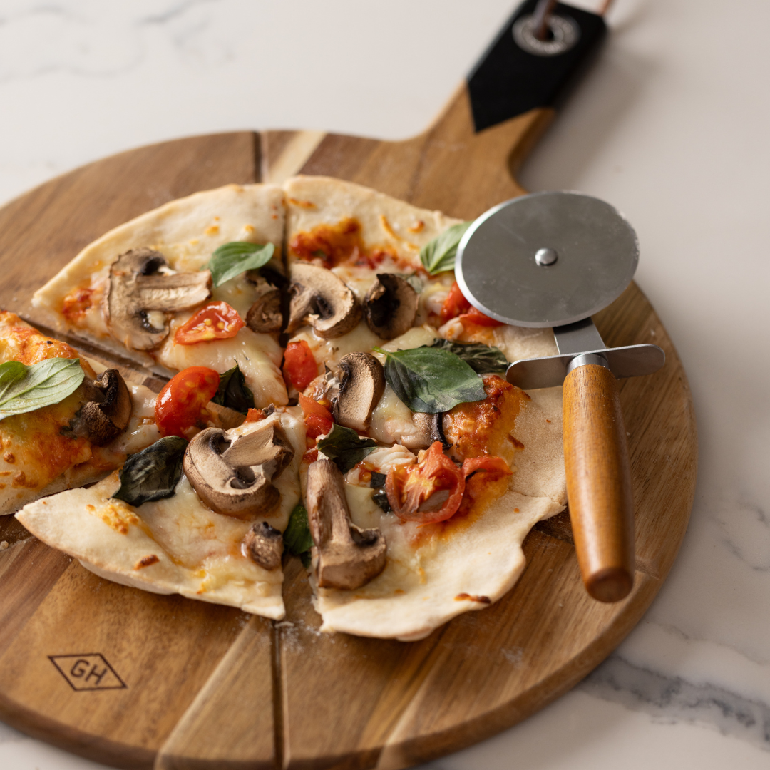 A partially sliced pizza with mushrooms, tomatoes, cheese, and basil rests on the Pizza Board with pizza cutter made of acacia wood.