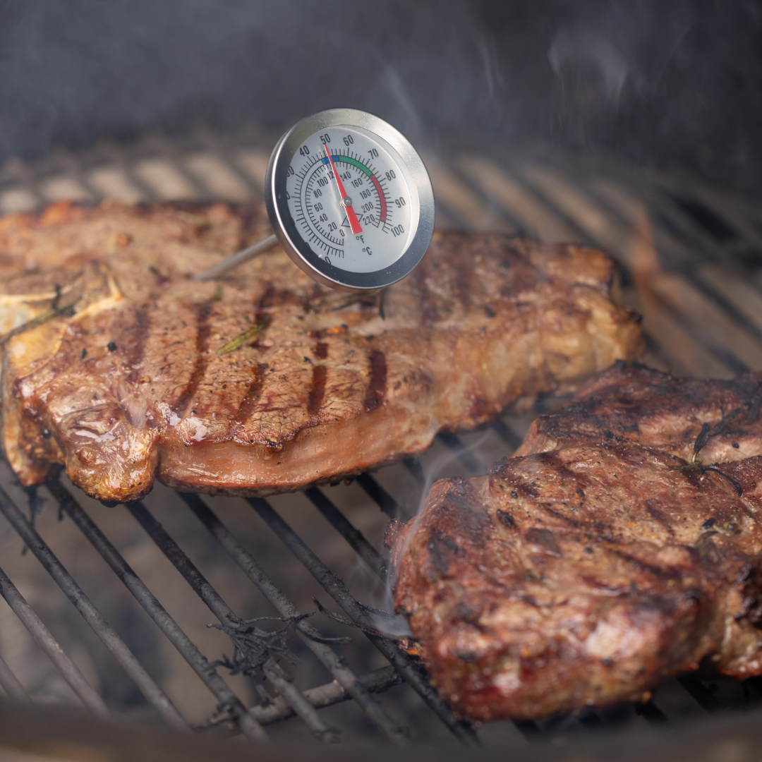 Two steaks sizzle on the grill, one tested with the BBQ Lovers Kit Meat Tenderizer & Thermometer as steam swirls—a perfect BBQ moment.