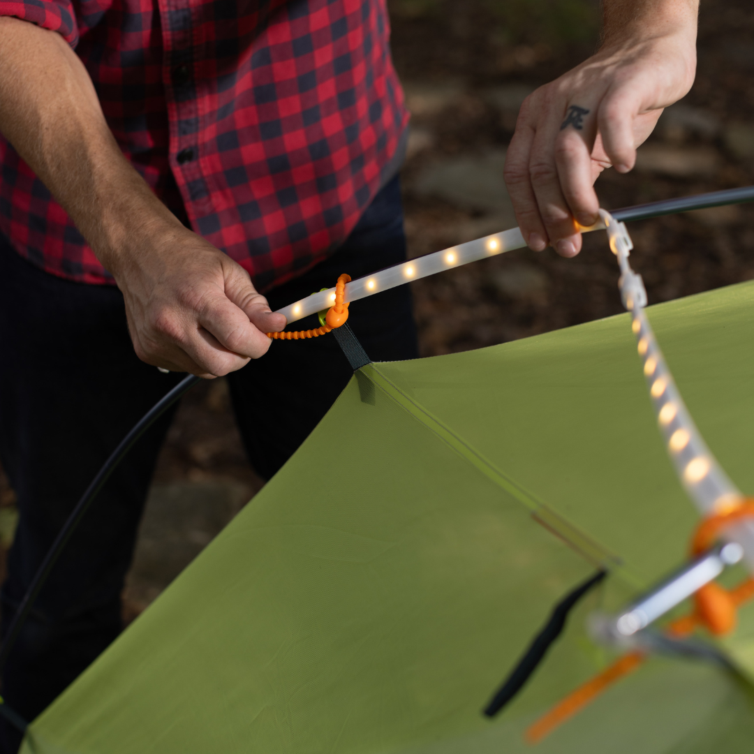 A person in a red plaid shirt attaches USB powered LED Rope Lights to the support poles of a green tent outdoors, enhancing their campsite lighting.