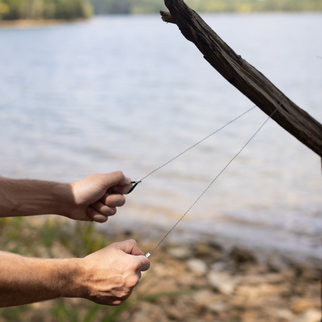 Hands using a wire saw to cut through a tree branch near a lakeshore—a must-have tool included in the Explorer Kit for outdoor adventures and exploring uncharted terrain.