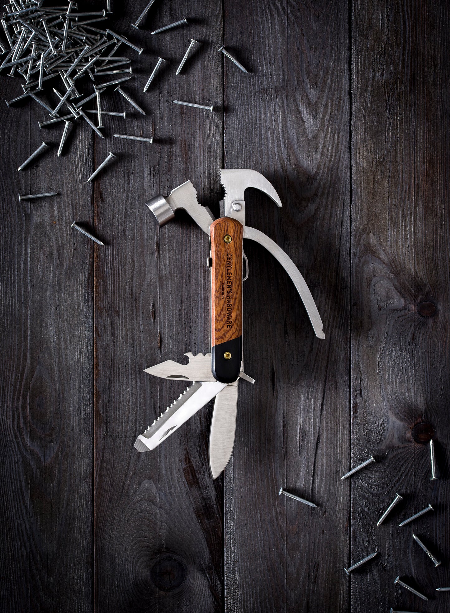 A Hammer Multi-Tool, Wood with stainless steel attachments—hammer, knife, and saw—rests on a dark wooden surface surrounded by scattered nails.