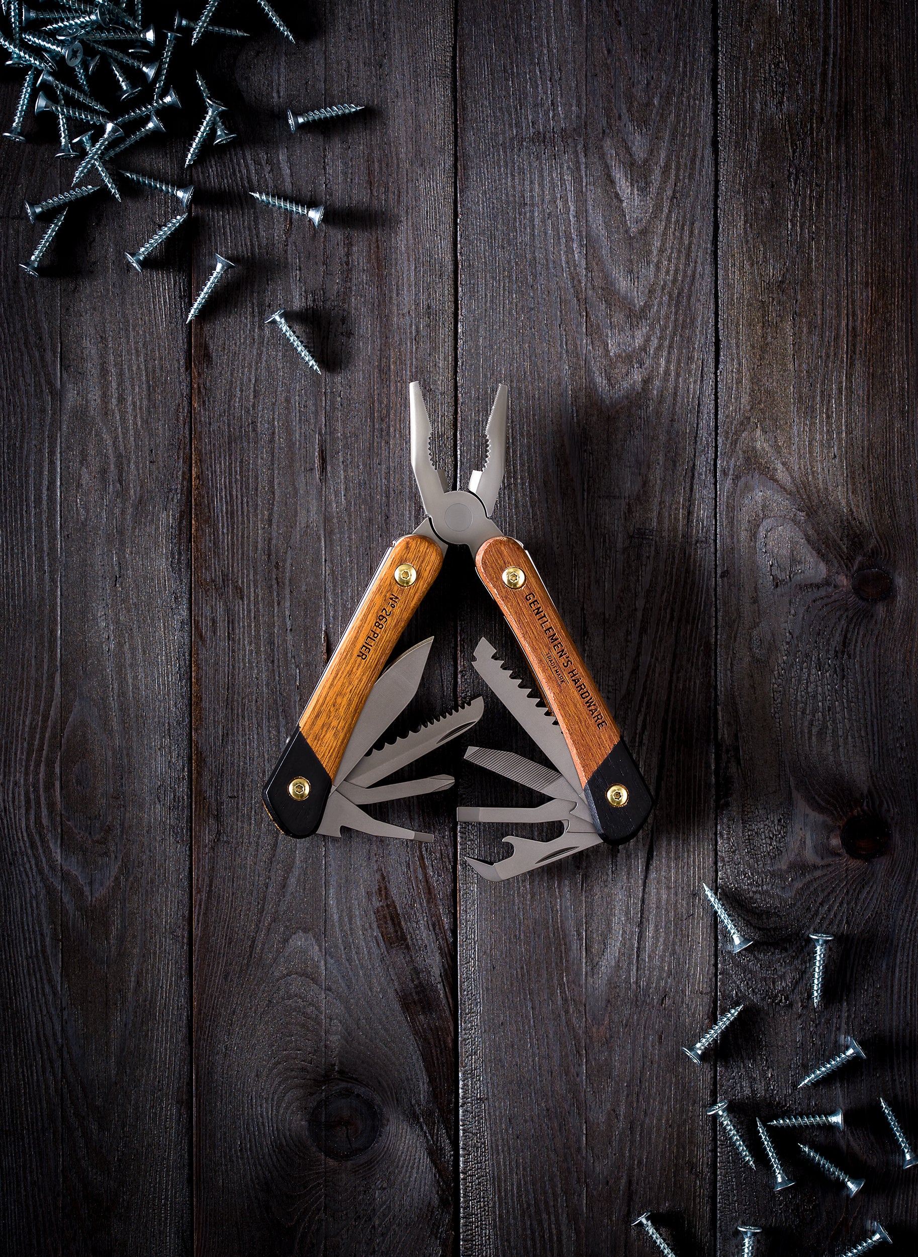 The Pliers Multi-Tool with wooden handles and open pliers, featuring a scratch-resistant finish, lies on a dark wooden surface amid scattered metal screws.