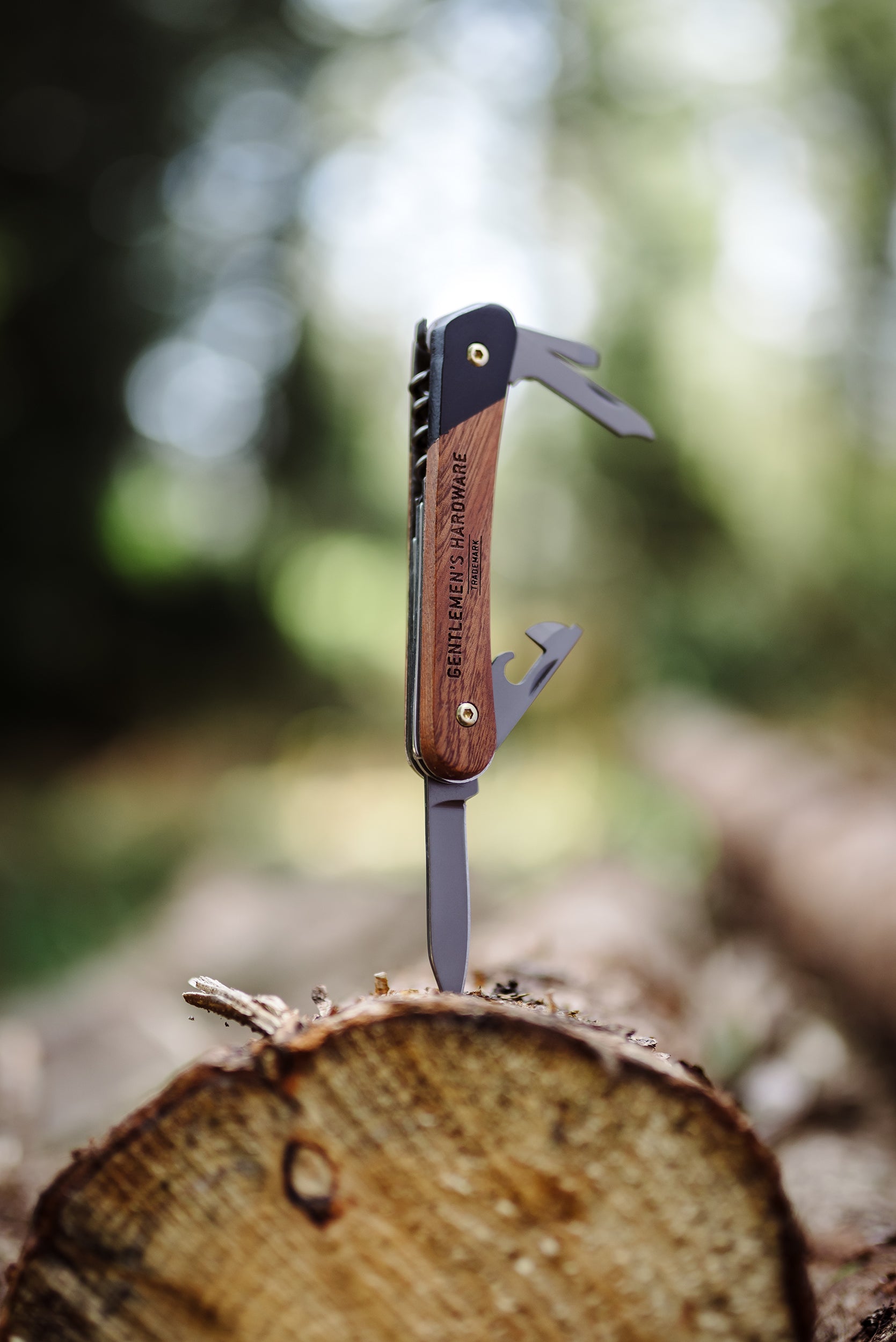 The Pen Knife Multi Tool, featuring a titanium coating and wooden handle, stands upright in a tree stump outdoors with blurred greenery in the background.