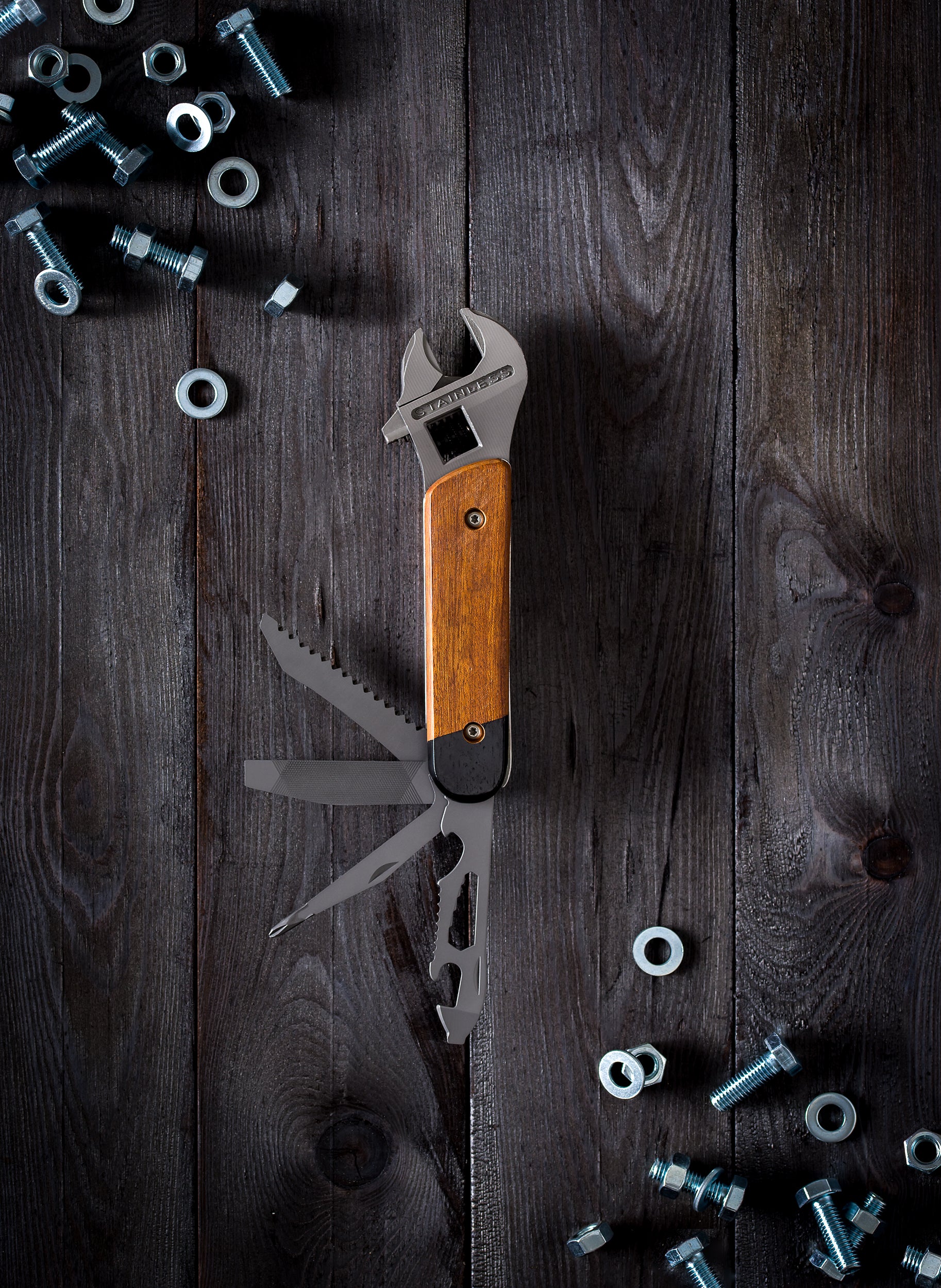 The Wrench Multi Tool, a heavy-duty multitool with a wooden handle and fold-out functions, rests on a dark wooden surface amid scattered bolts and nuts.