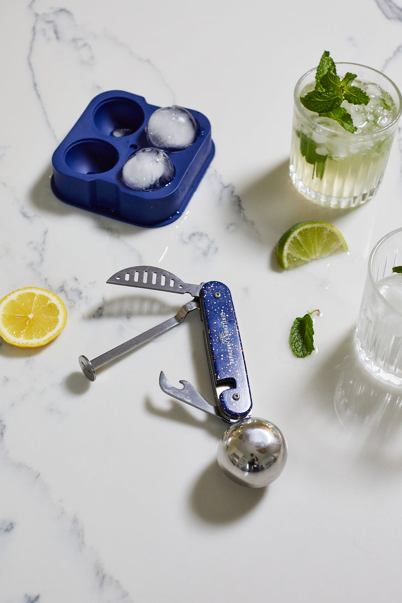 A Cocktail Multi-Tool, a blue ice mold with round ice balls, sliced lemon and lime, and two mint-garnished drinks are arranged on a white marble surface.