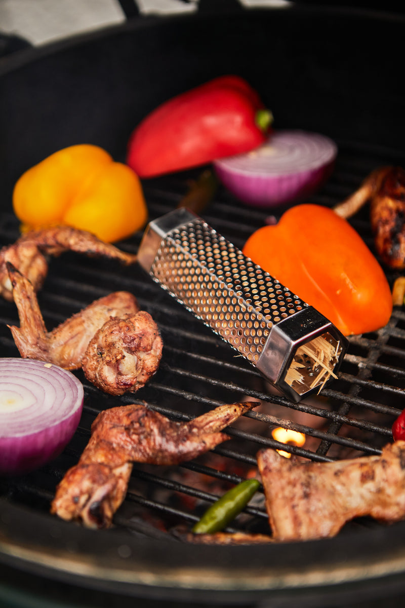 A grill with chicken wings, red and yellow bell peppers, red onion halves, and a BBQ Smoker Kit placed on the grates as a flavorful grilling accessory.