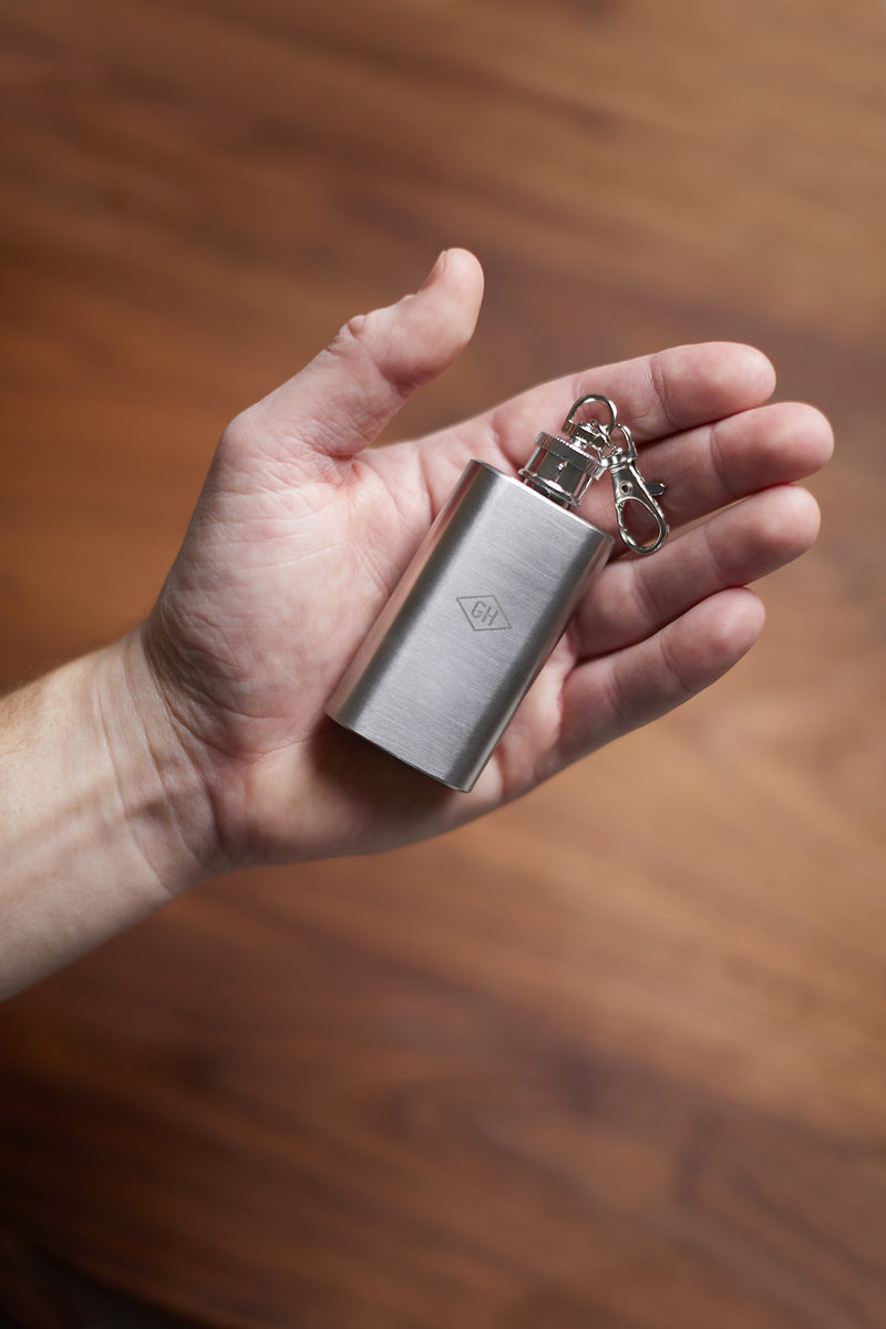 A hand holds the Mini Hip Flask keychain, crafted from stainless steel, against a wooden background.