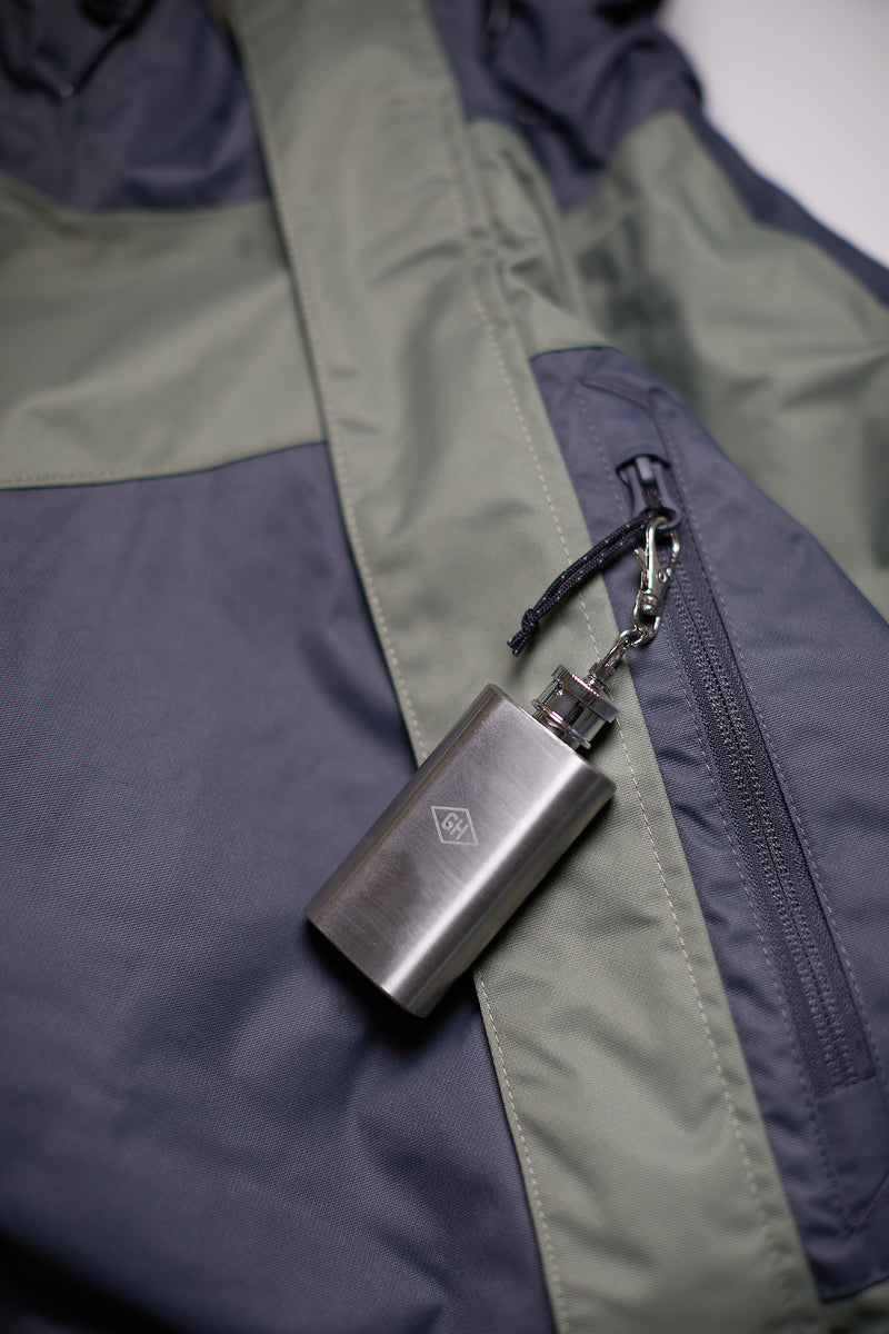 A Mini Hip Flask made of stainless steel is attached to a zipper pull on a two-tone green and gray jacket.