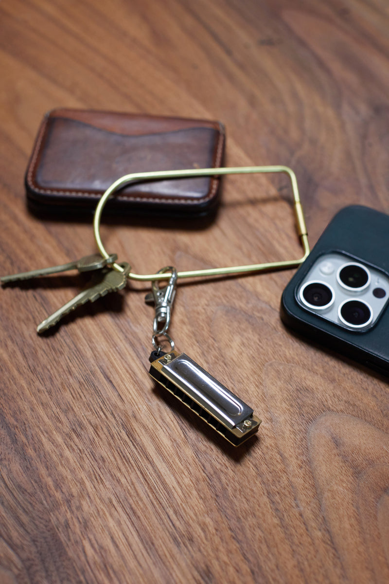 A set of keys, a Mini Harmonica keychain, a leather wallet, and a smartphone with triple cameras rest on a wooden surface.