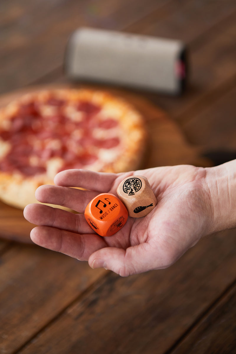 A hand holds two Friday Night Dice, one orange and one wooden, displaying icons and text. A blurred pepperoni pizza sits in the background—making Friday Night Dice an ideal, unique gift for fun takeaway decisions.