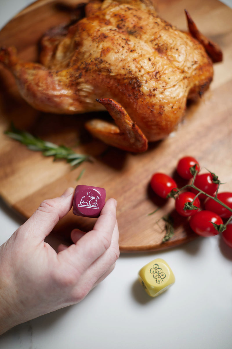 A roasted chicken on a wooden board with rosemary and vine tomatoes; a hand holds the burgundy BBQ Dice with a food icon, while the yellow dice featuring a bee icon adds fun to this BBQ Dice grilling game scene.