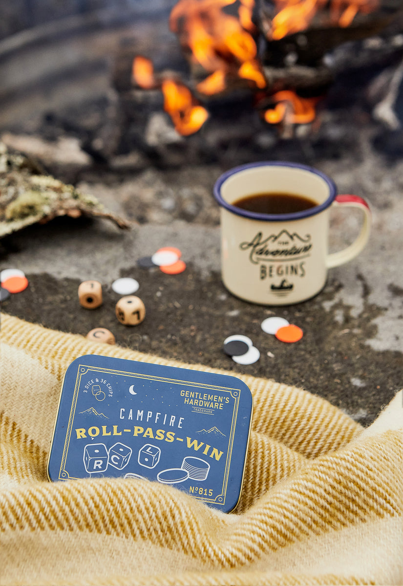 A tin of Campfire Roll Pass Win travel game sits by a campfire, alongside a mug of coffee, wooden dice, and game pieces on a blanket.