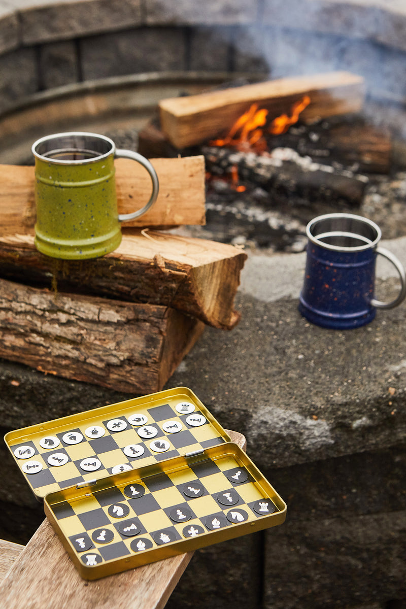 The Travel Chess set with magnetic pieces and two metal mugs are placed on firewood and stone near glowing campfire logs.