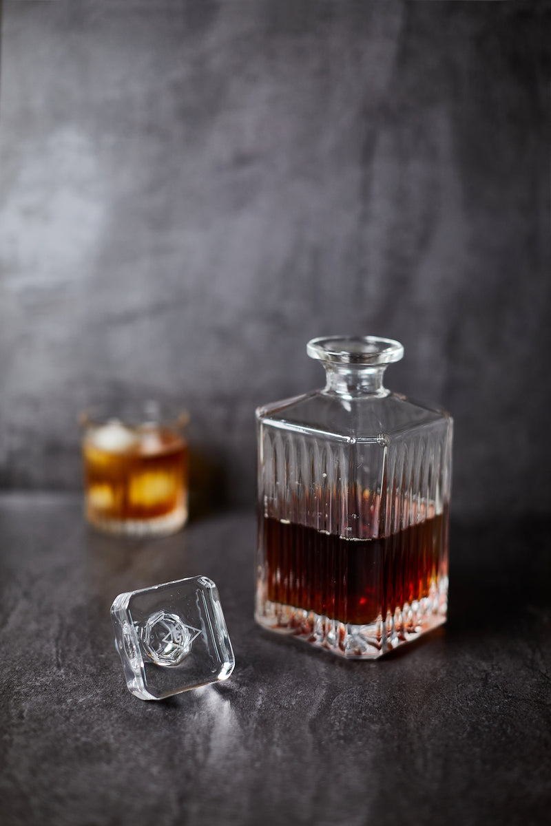 A Whisky Decanter filled with dark liquor rests on a dark surface beside its stopper, with a glass of whiskey and ice in the background—ideal for enhancing your at-home bar collection.