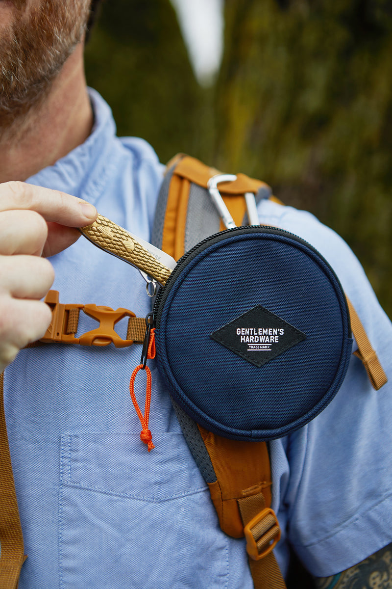 A person wearing a blue shirt and brown backpack holds a small tool next to a round navy Utility Pouch labeled Gentlemens Hardware, which is attached to the backpack strap with a carabiner.
