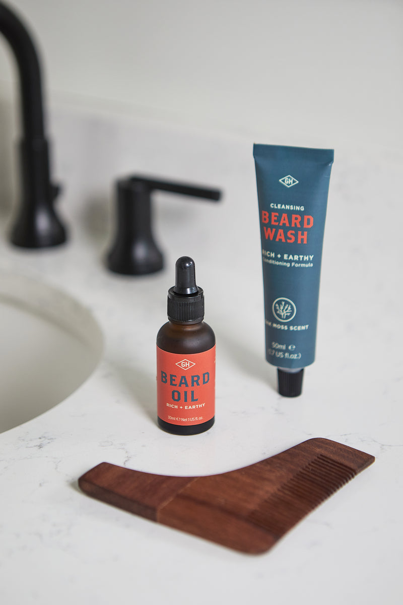 The Beard Survival Kit, which includes Beard Oil, Beard Wash, and a wooden comb, is neatly arranged on a bathroom counter near the sink and black faucet.