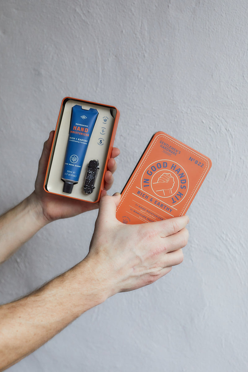 A person holds an open rectangular tin box containing the In Good Hands Kit—featuring a hand moisturizer and manicure multi-tool—against a light gray textured background.