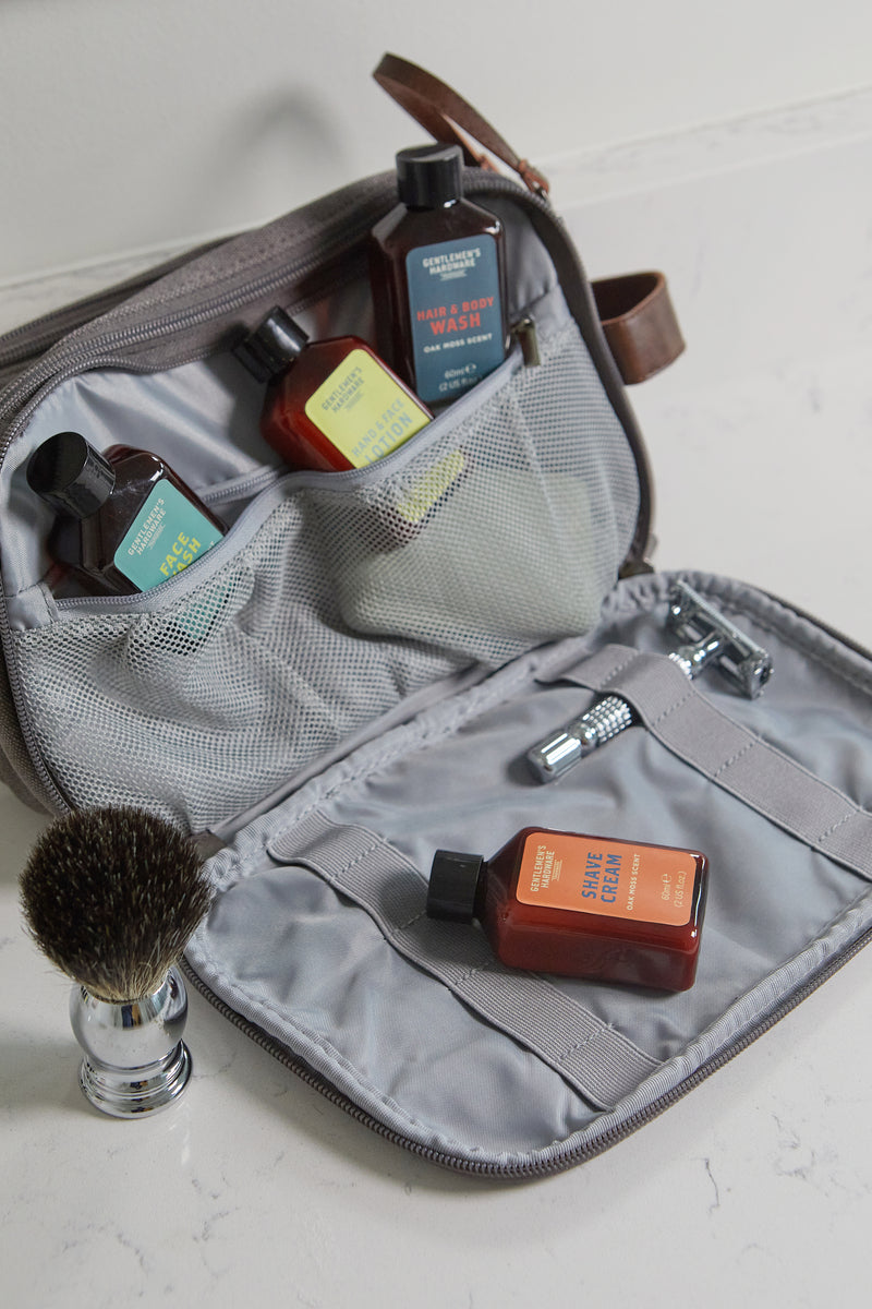 The Getaway Grooming Kit features a gray toiletry bag with mesh pockets for small personal care bottles. A shaving brush and razor sit nearby on a white surface, making it the perfect compact travel accessory.
