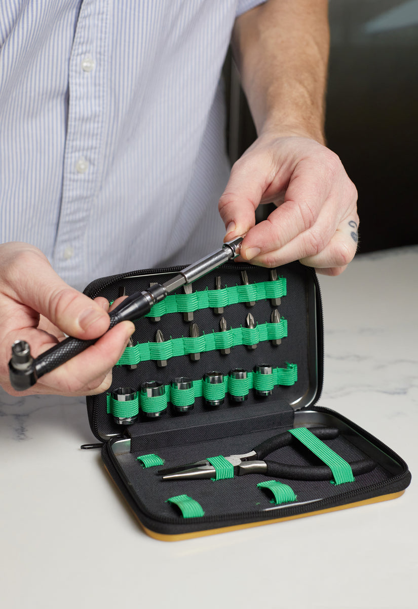 A person holds a tool from a black zippered Mini Tool Kit case, which contains assorted sockets, bits, and a ratchet—all organized with green elastic bands.