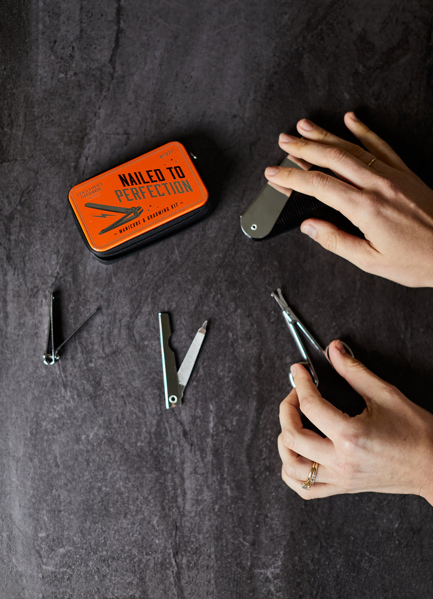 Someone uses a Manicure Kit nail clipper labeled Nailed to Perfection on a dark surface, with additional grooming tools arranged nearby.