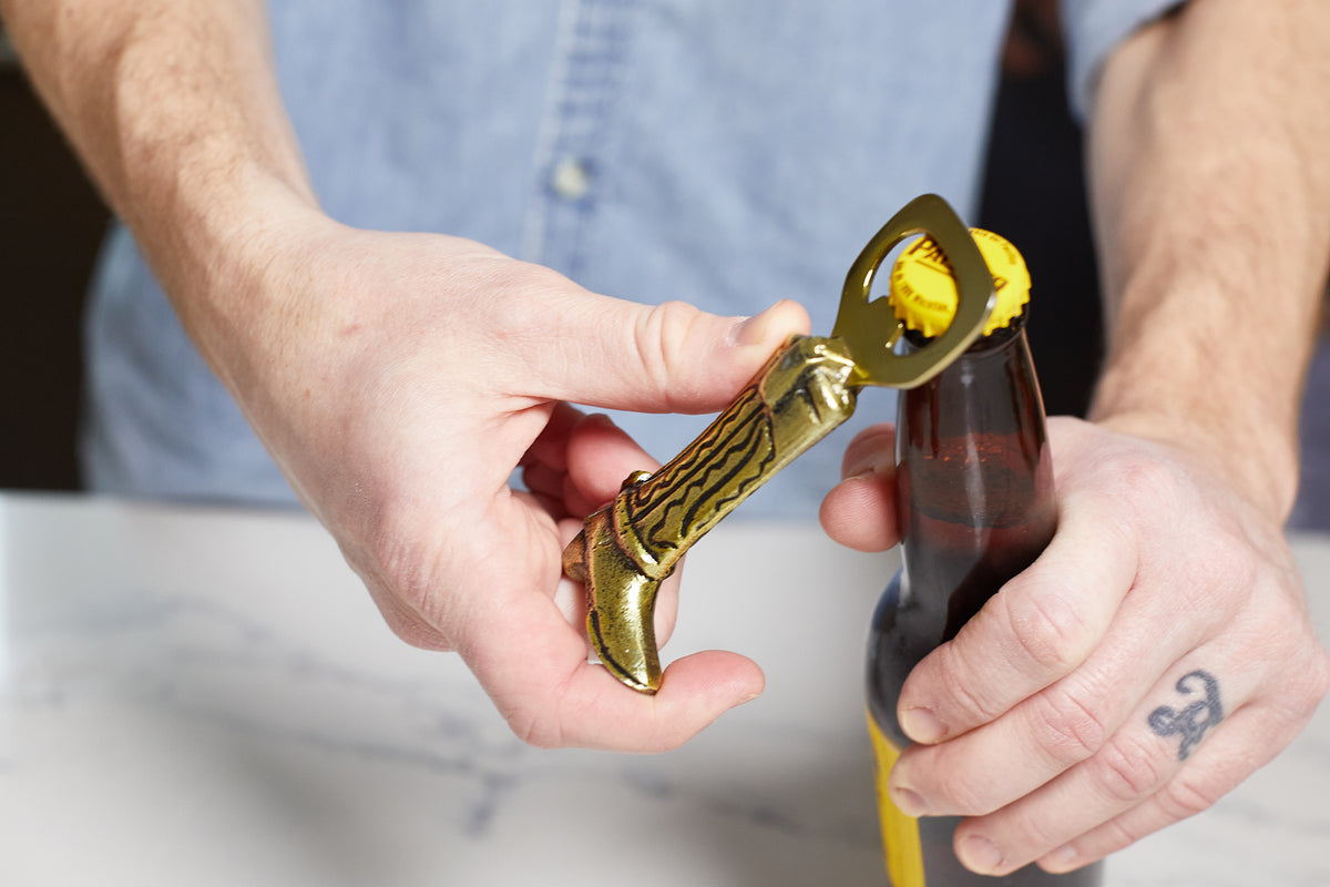 A person uses the Cowboy Boot Bottle Opener, featuring a gold-colored brass effect finish and shaped like a cowboy boot, to open a brown bottle—a striking bar accessory.