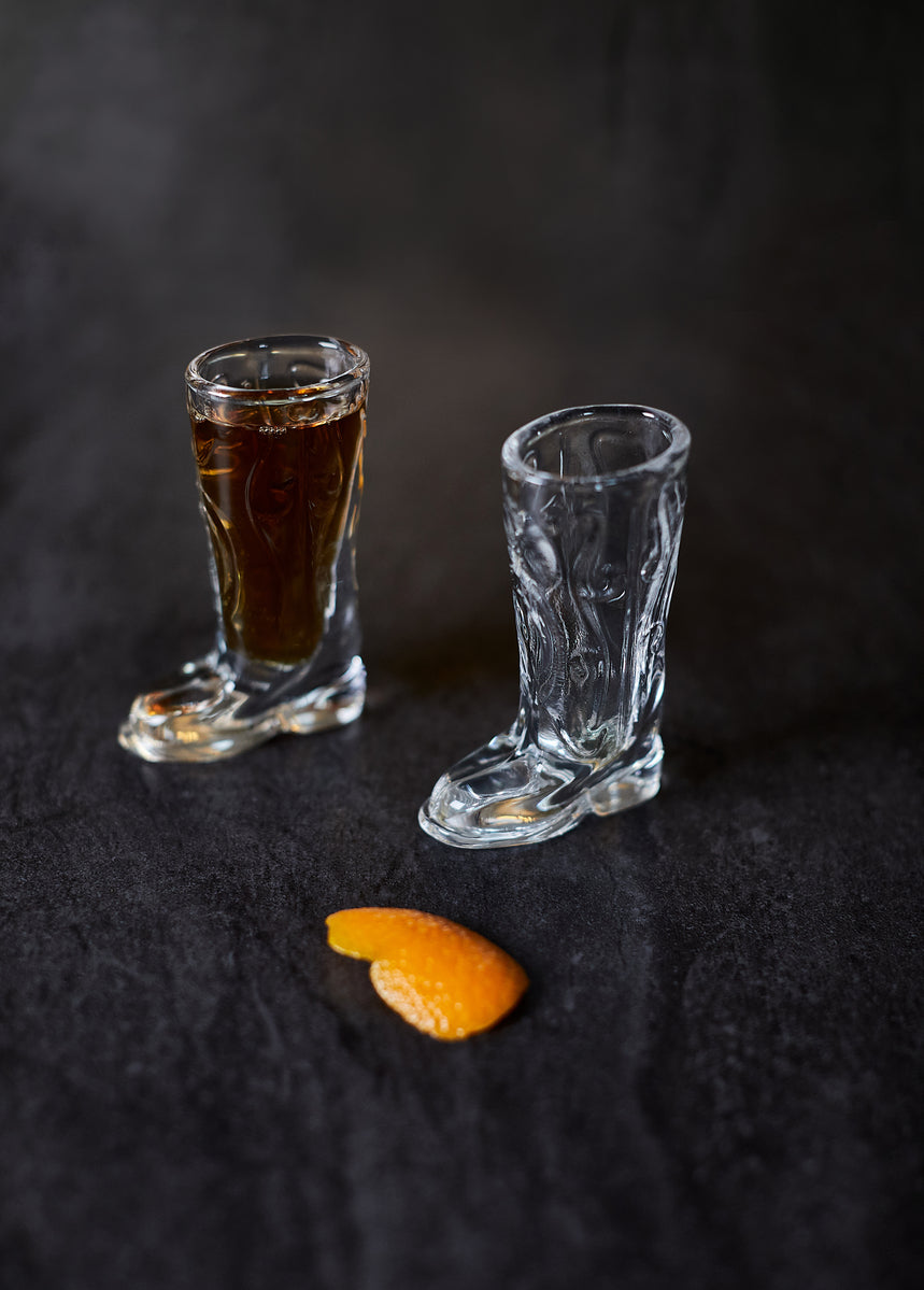Two shot glasses from the Set of 2 Cowboy Boot Shot Glasses—one filled, one empty—sit on a dark surface beside orange peel. A perfect addition to any at-home bar kit from Gentlemens Hardware.