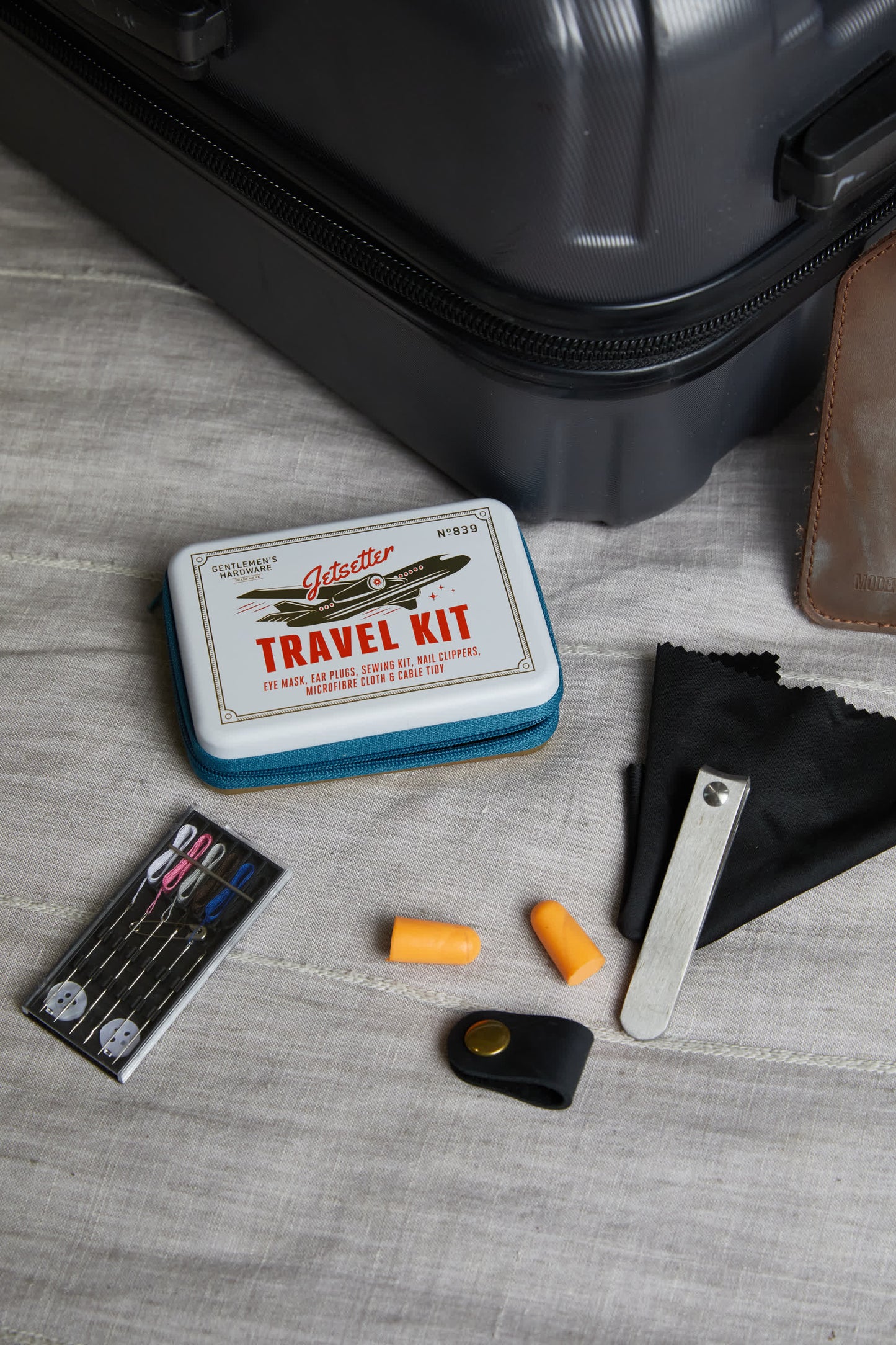 The Jet Setter Kit, a compact travel set with sewing needles, earplugs, a nail file, and a cleaning cloth, is displayed beside a suitcase on light fabric—ideal for keeping essentials handy on the go.